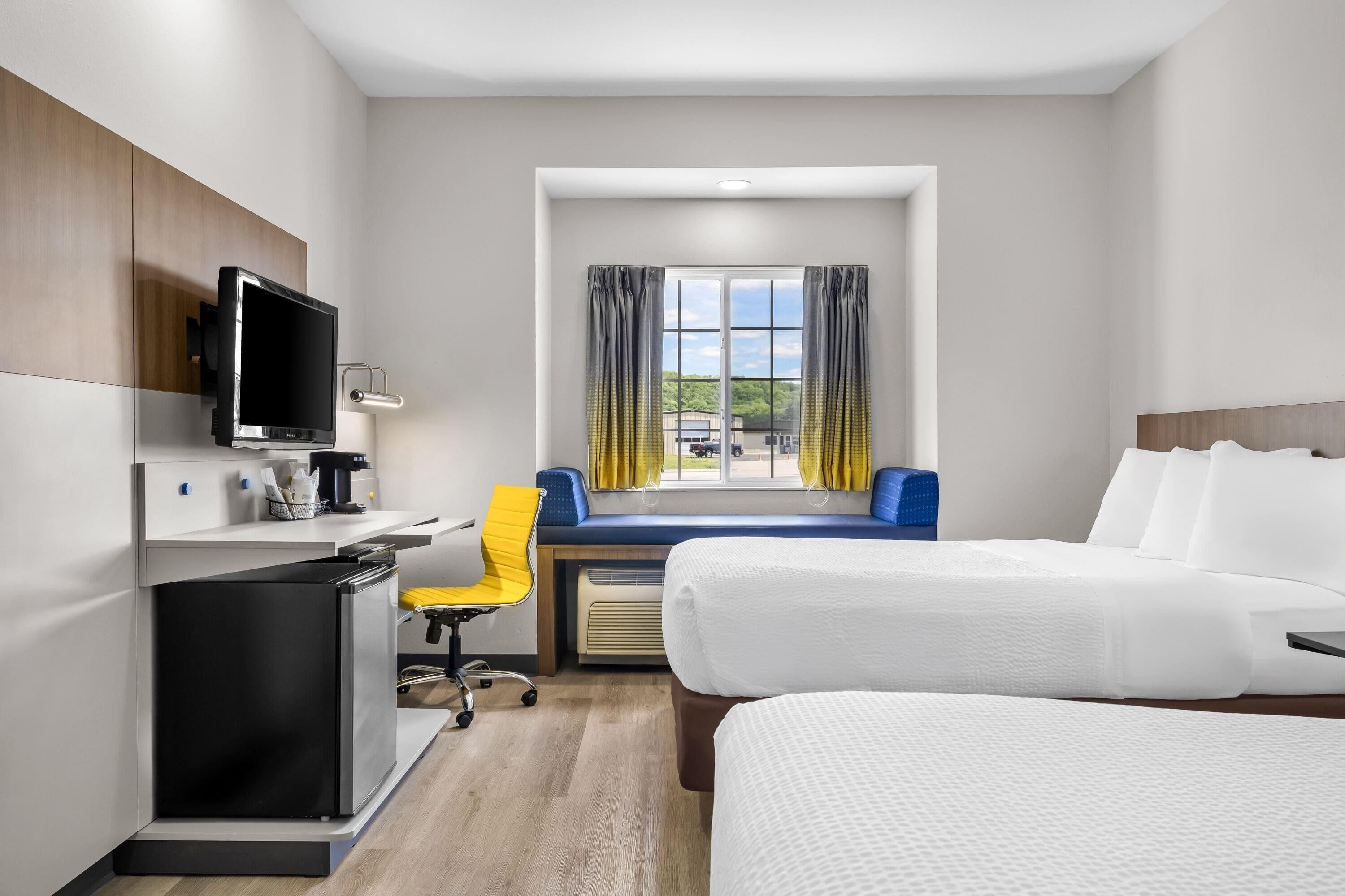 Una habitación de hotel con cama, escritorio, silla, televisor y ventana con cortinas.