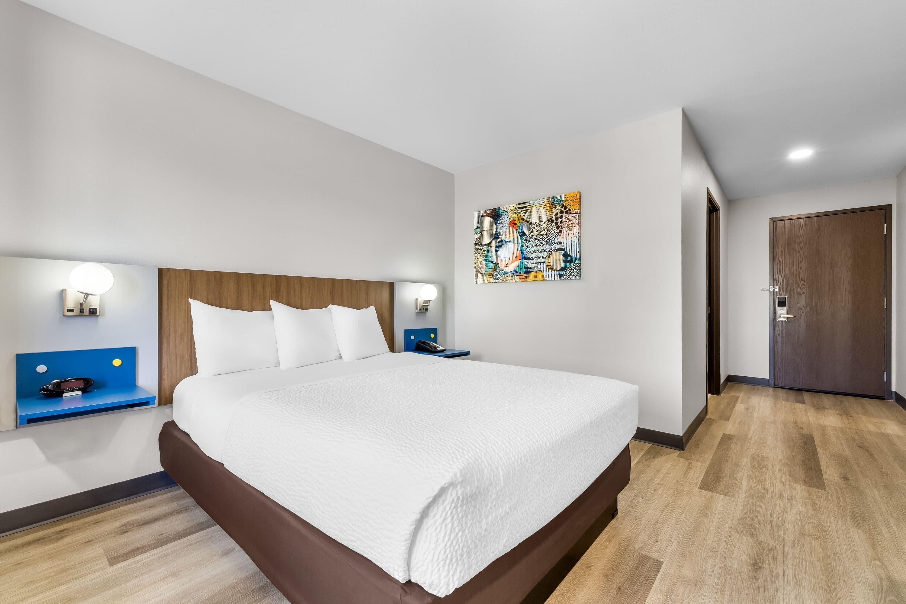 A modern hotel room with a large bed, bedside tables, a wall-mounted artwork, and a wooden door.