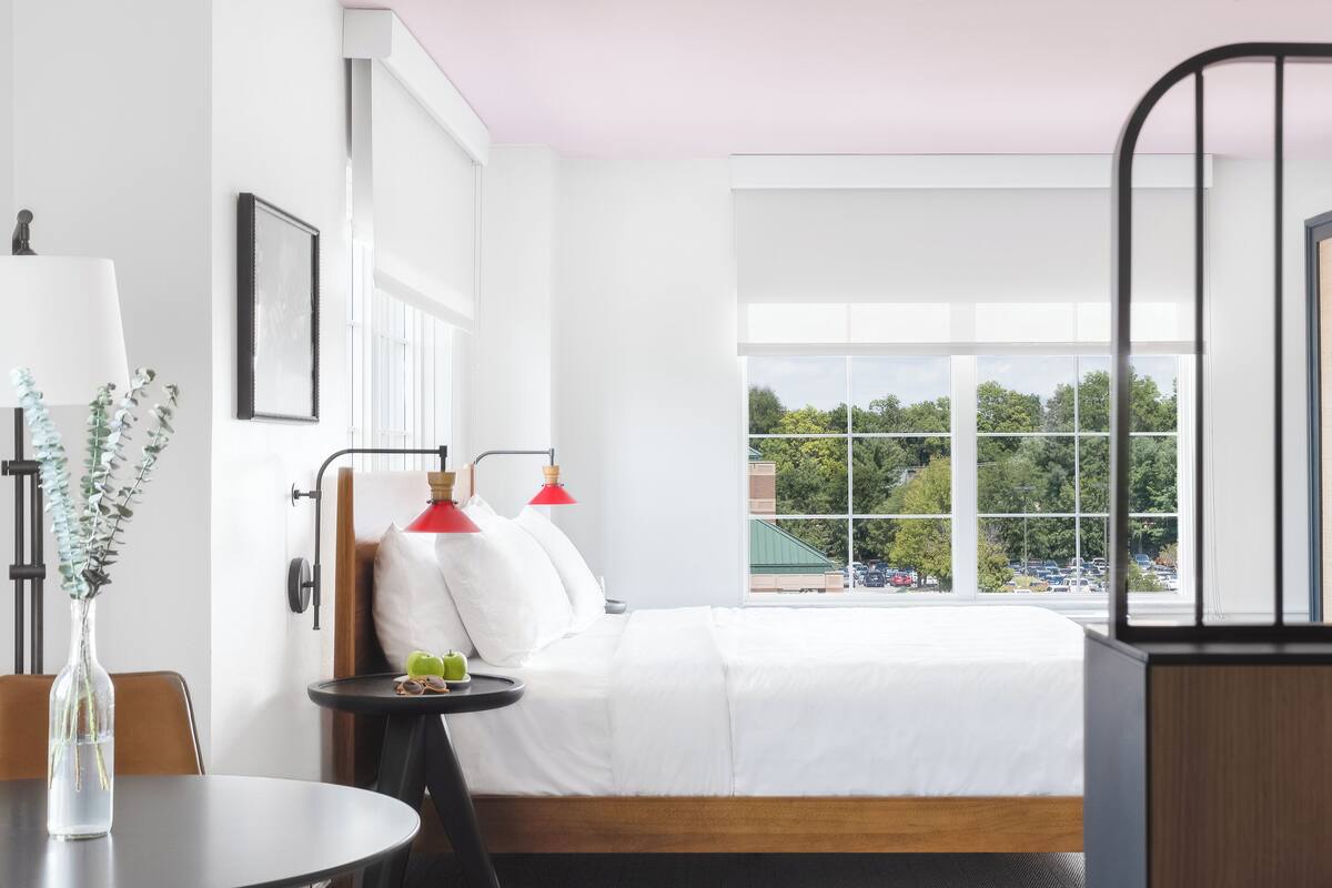 Ein Hotelzimmer mit großem Fenster, einem Bett mit weißer Bettwäsche, einem kleinen Tisch mit einer Vase und roten Lampen.