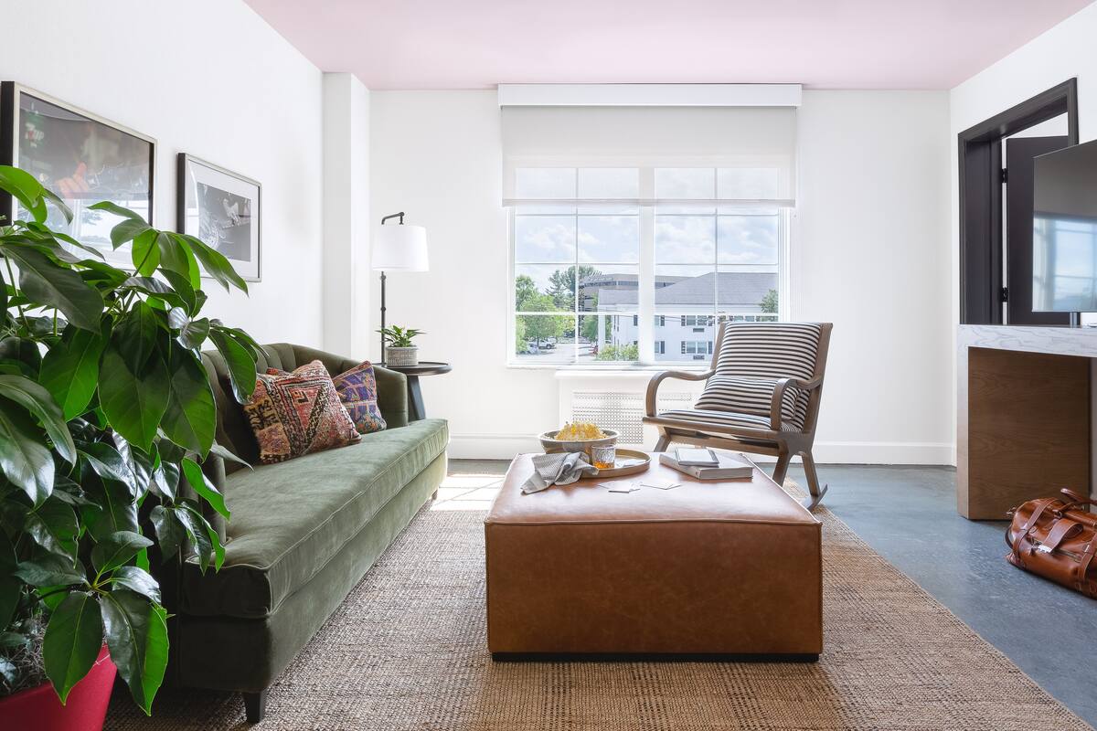 Ein Wohnzimmer mit einem grünen Sofa, einem gestreiften Sessel, einem braunen Hocker und einem großen Fenster mit Blick auf Gebäude.