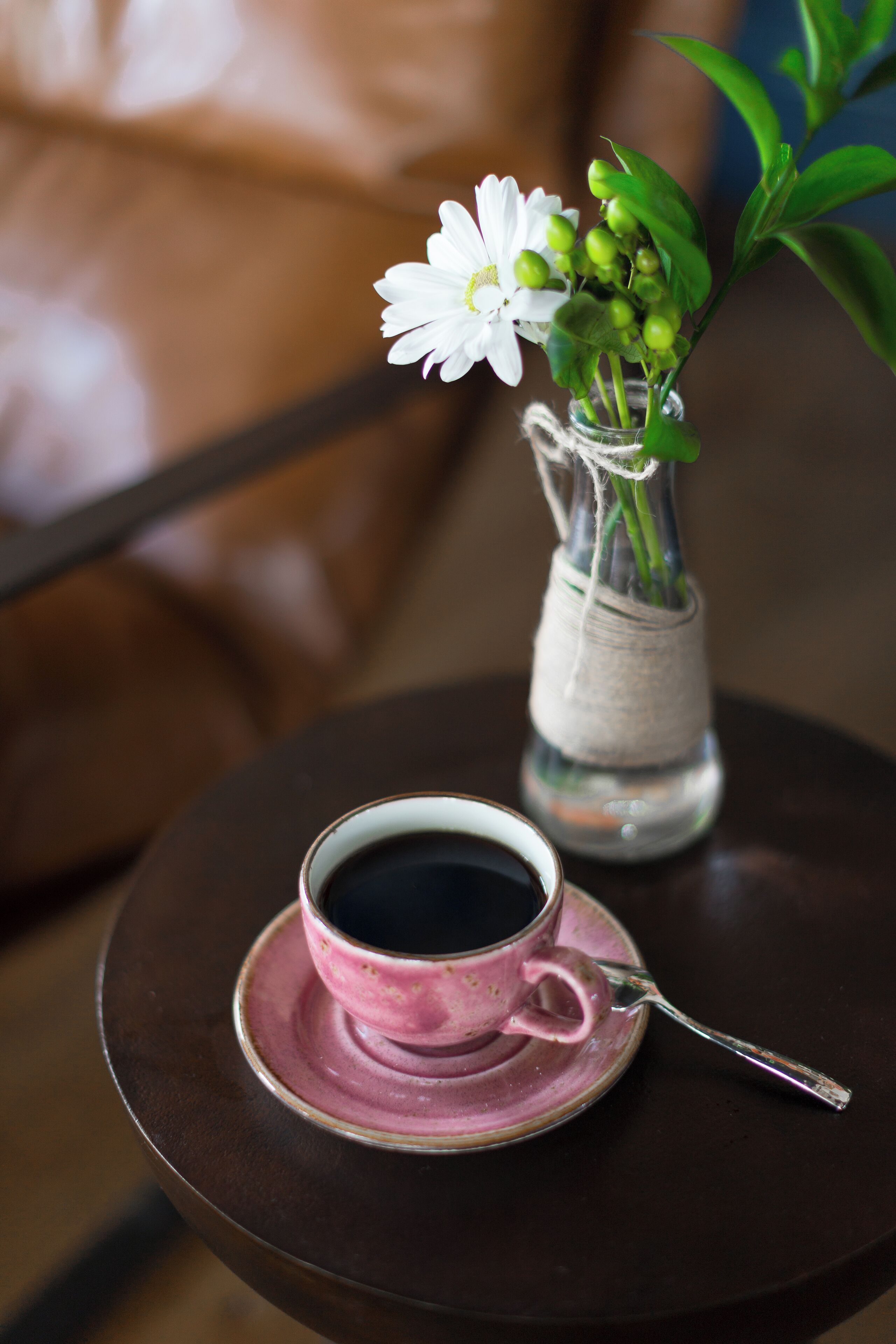 A cup of coffee on a saucer with a spoon, a small vase with a flower, and a brown leather chair in the background.