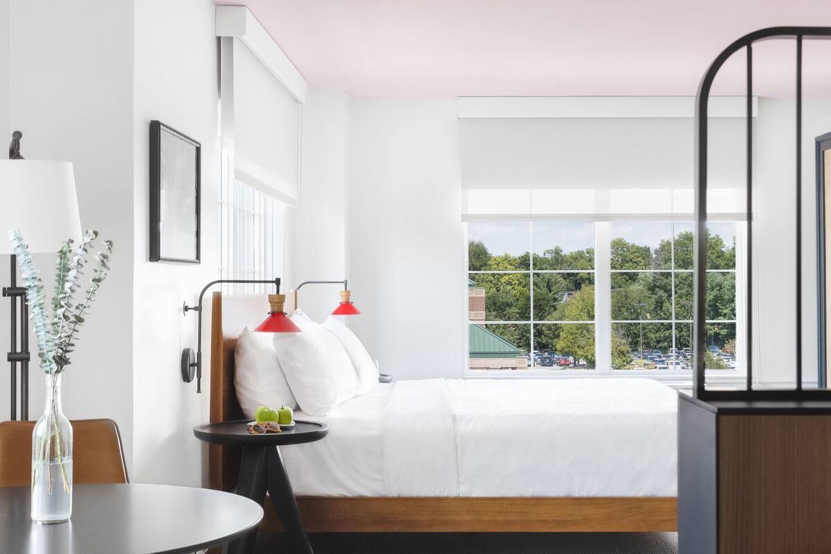 Ein modernes Hotelzimmer mit einem großen Bett, zwei Nachttischlampen, einem kleinen Tisch mit einer Vase und Blick auf einen Parkplatz durch große Fenster.