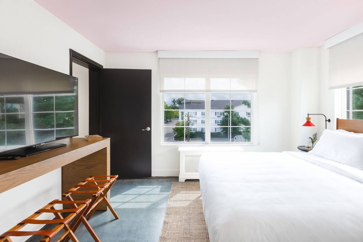 Ein modernes Schlafzimmer mit Bett, Schreibtisch mit Laptop, Stuhl, Fenster mit Blick auf Gebäude und einer roten Lampe.