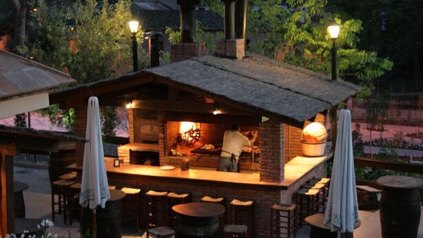 Un área de bar al aire libre con una chimenea de ladrillo, taburetes de madera y un chef preparando comida.