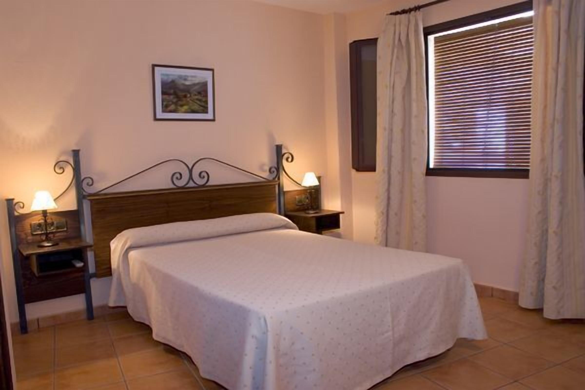Una cama individual en habitación con cabecera de madera, dos mesitas de noche con lámparas, un cuadro enmarcado en la pared y una ventana con cortinas.