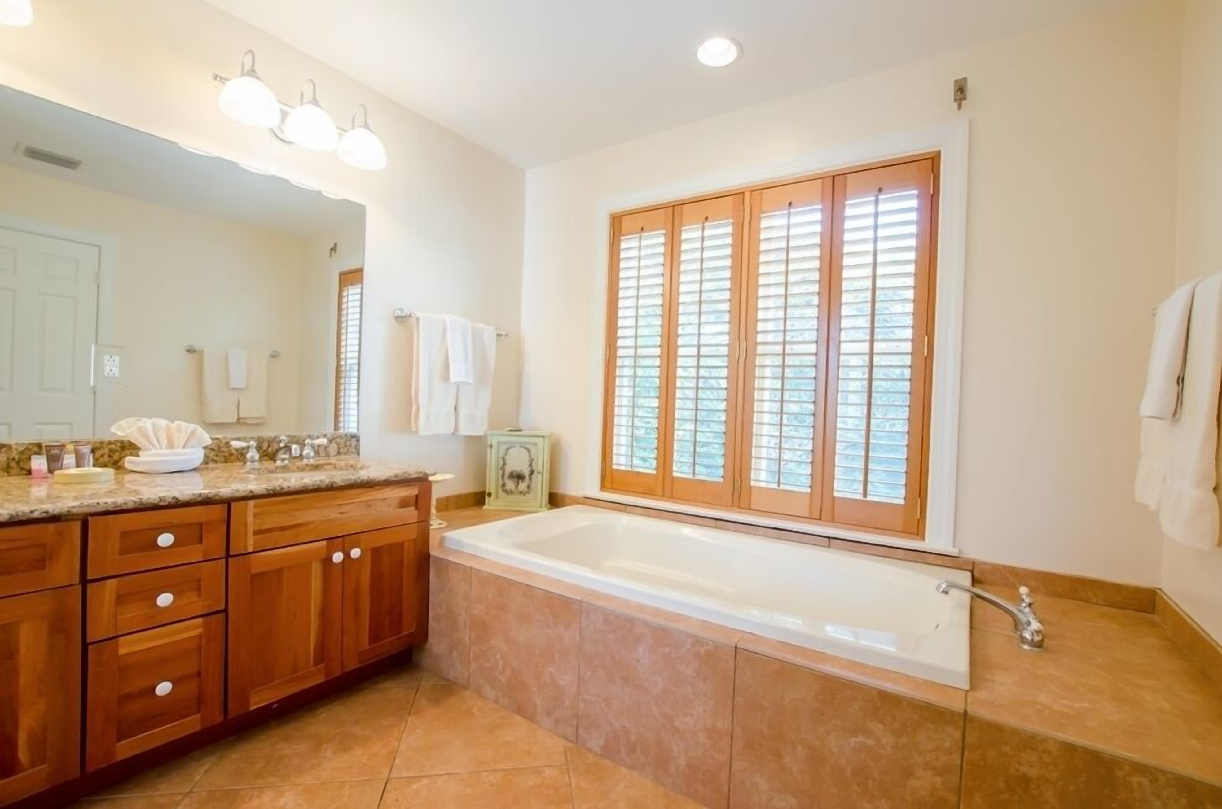 A bathroom with a large bathtub, wooden cabinets, a vanity with a mirror, and a window with blinds.