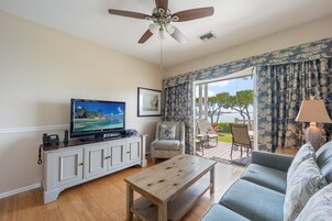 Uma sala de estar com televisão, ventilador de teto, sofá, mesa de centro e vista para o exterior através de grandes janelas.