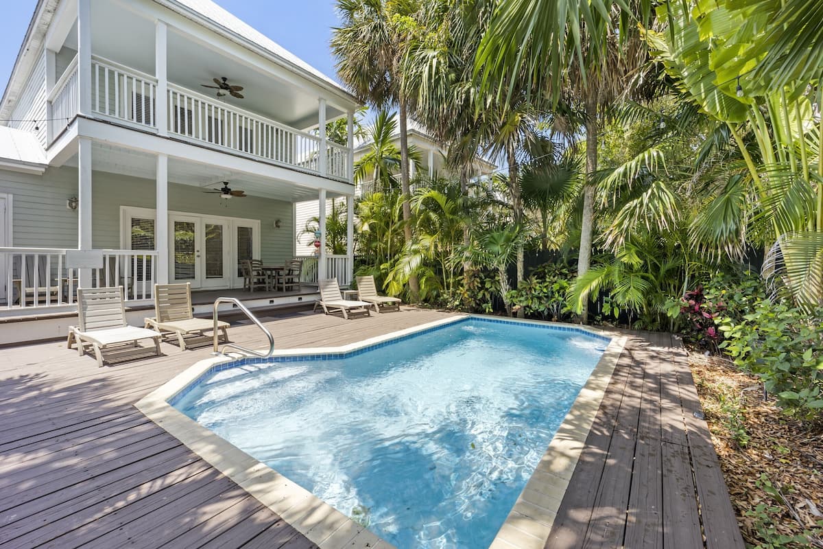 Ein Pool mit Holzterrasse und Liegestühlen, umgeben von tropischen Pflanzen und einem zweistöckigen Haus mit Balkonen.