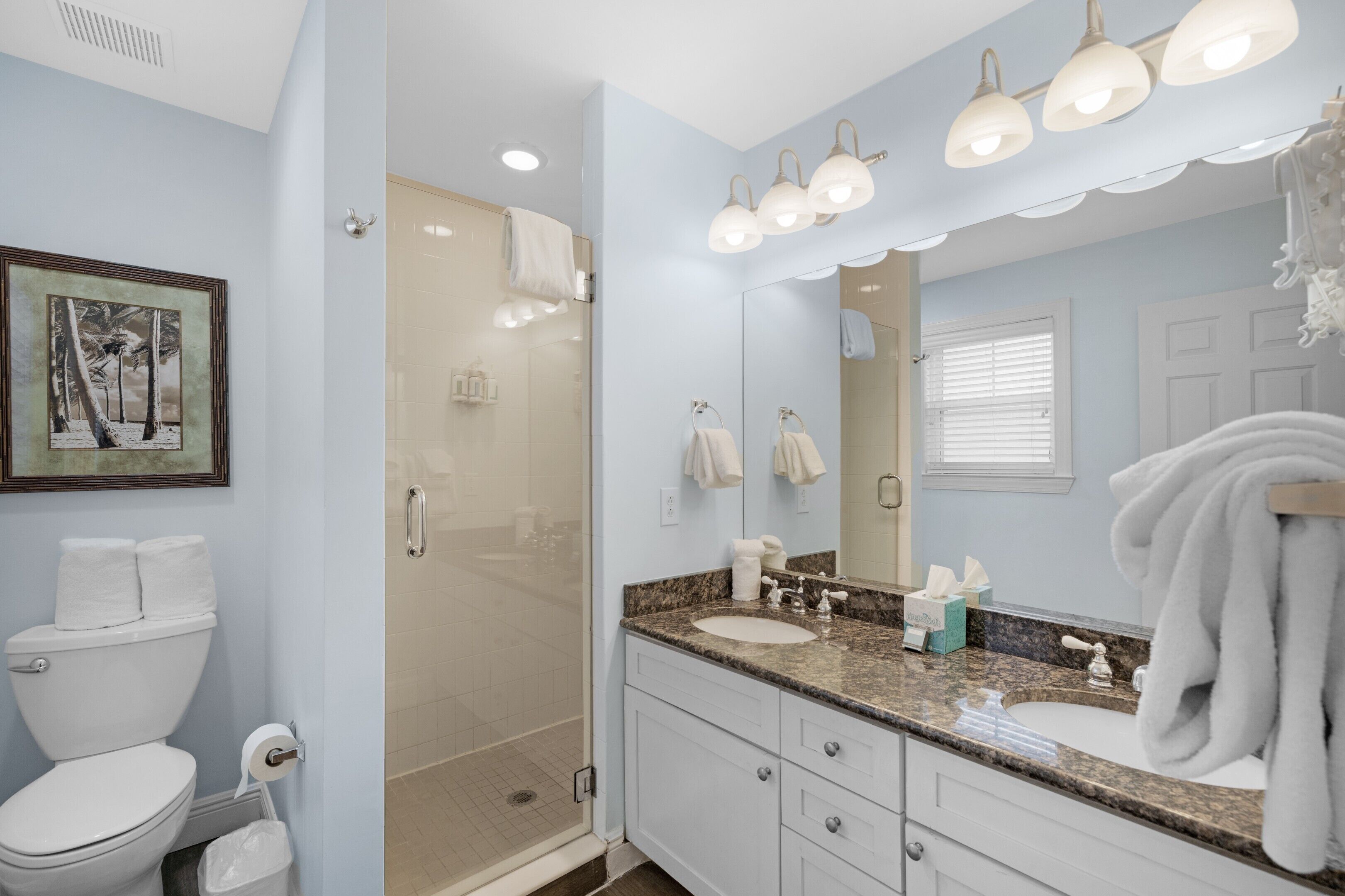 A bathroom with a glass-enclosed shower, a sink with a vanity, a large mirror, and a framed picture on the wall.
