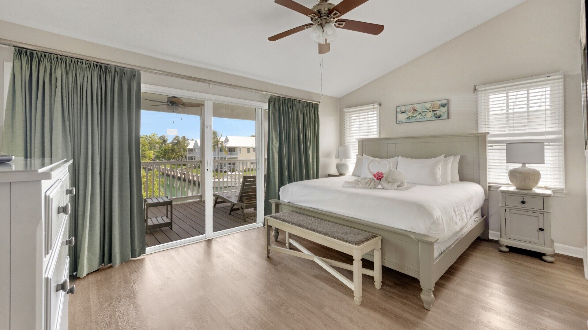 A bedroom with a large bed, a bench, a nightstand, a ceiling fan, and a view of the outside.