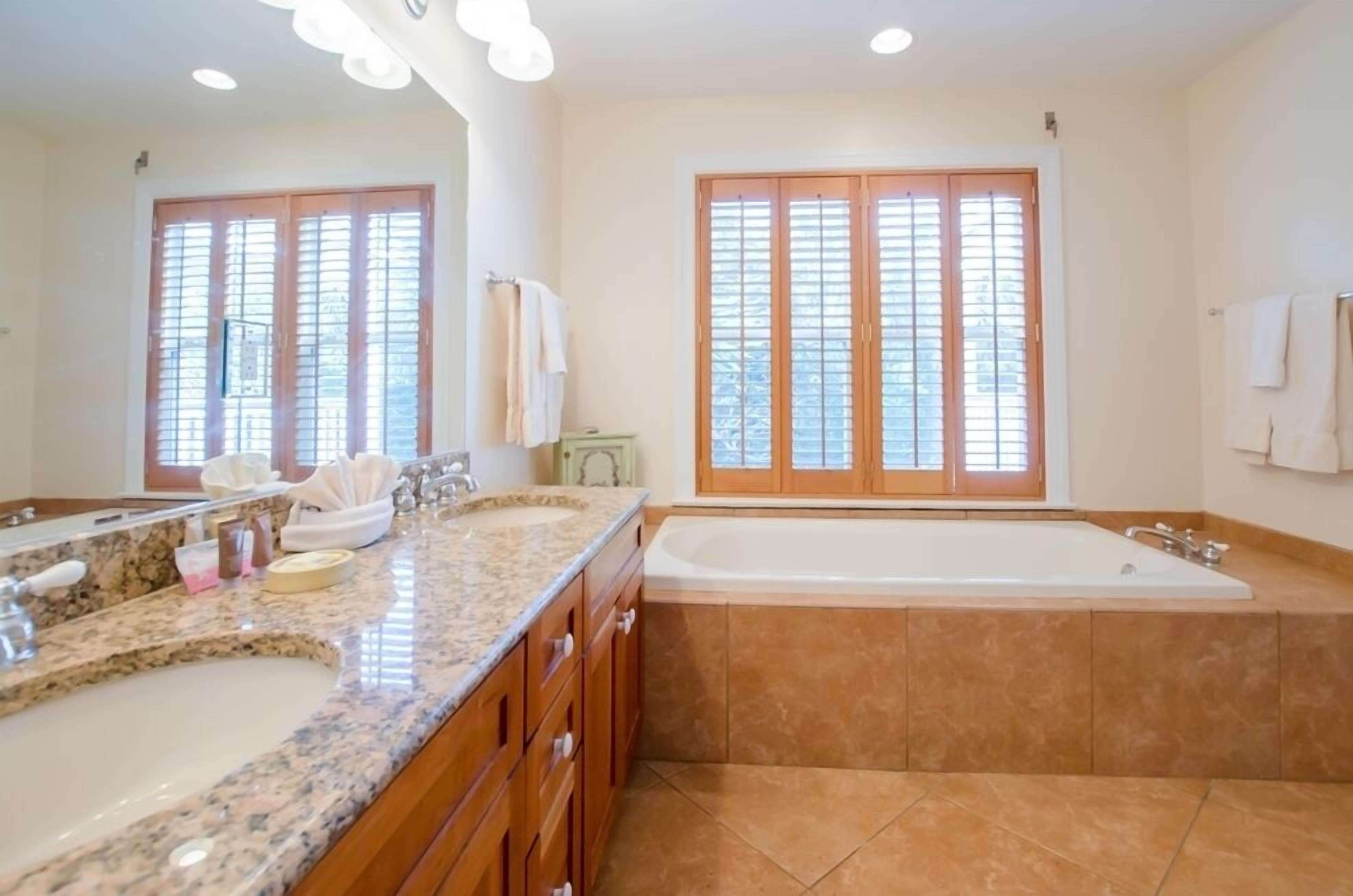 A bathroom with two sinks, a bathtub, and large windows with blinds.