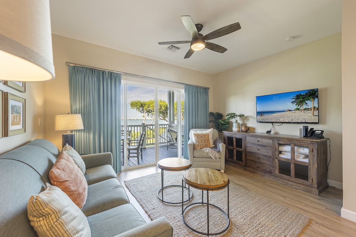 Ein Wohnzimmer mit einer Couch, zwei runden Tischen, einem Fernseher und einem Balkon mit Blick auf den Strand.