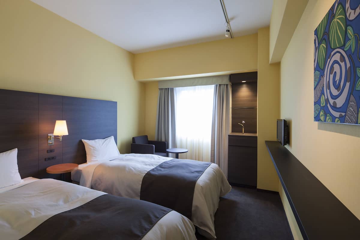 Hotellihuone, jossa on kaksi sänkyä, pieni pyöreä pöytä, työpöytä ja suuri ikkuna, jossa on verhot.