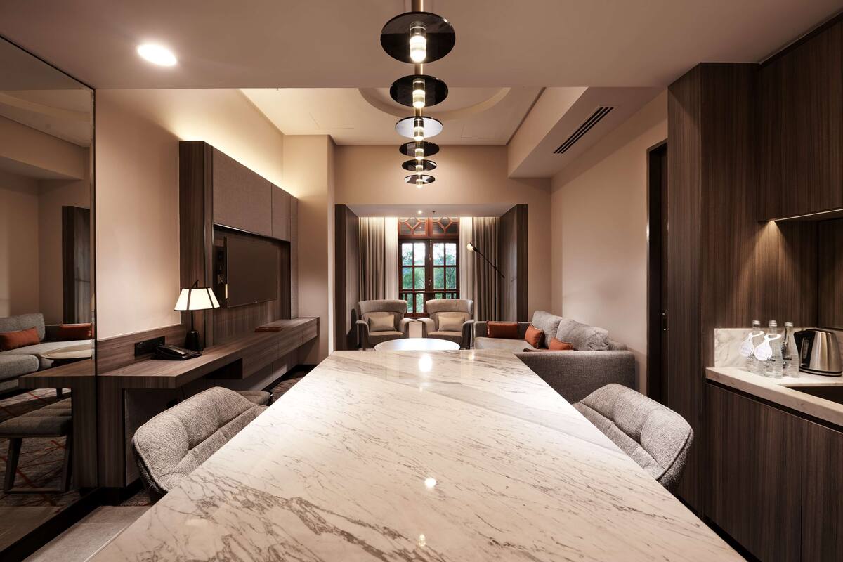 A modern hotel room with a marble dining table, two sofas, a TV, and a minibar.