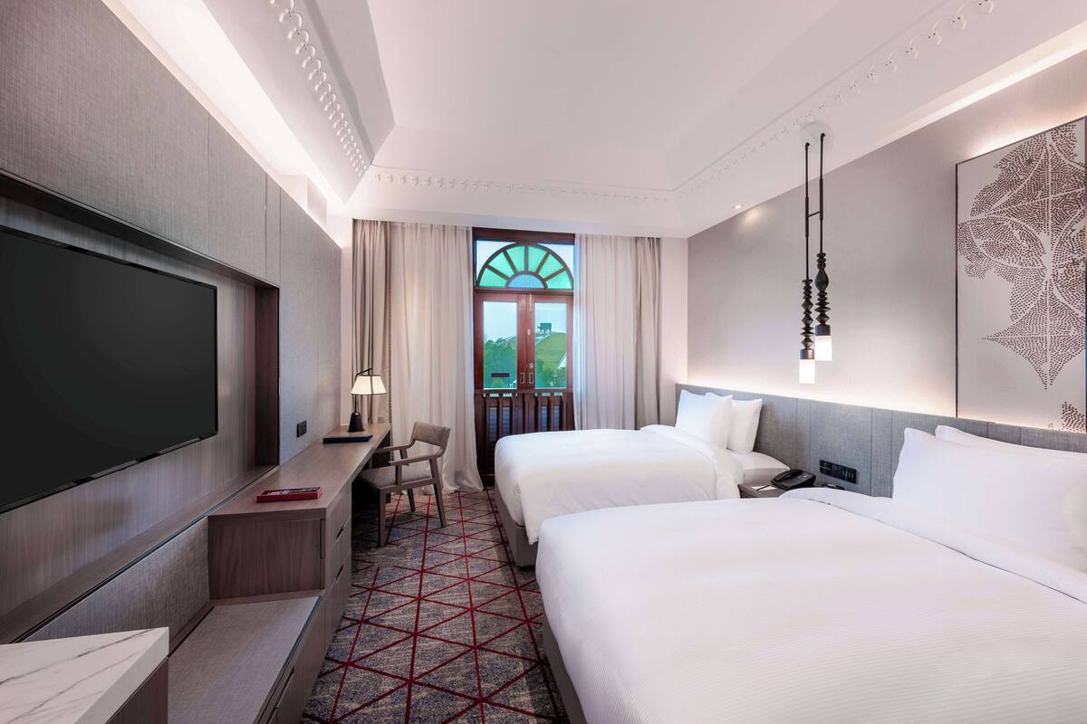 A hotel room with two beds, a TV, a desk, and a window with a view of a green landscape.