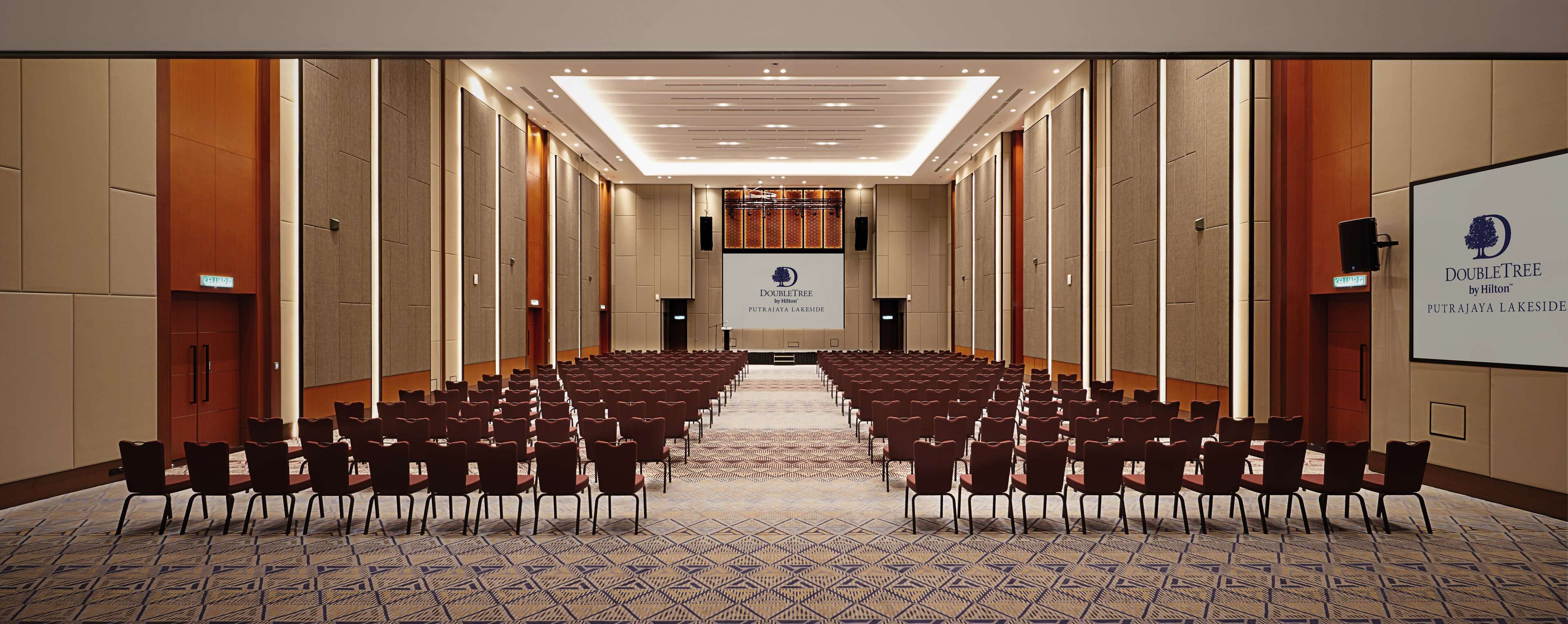 A conference room with rows of chairs facing a projection screen, a carpeted floor, and a wooden panel wall.