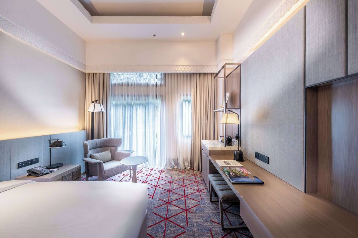 A modern hotel room with a large bed, a desk, a chair, and a small table. There is a window with curtains, a lamp, and a shelf with items on it.