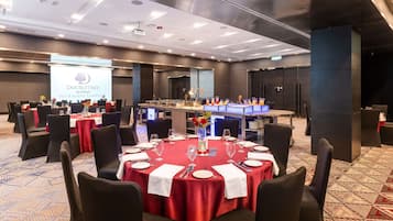 Sala de conferências com mesas redondas, toalhas vermelhas e cadeiras pretas. Uma tela de projetor exibe o logotipo DoubleTree.