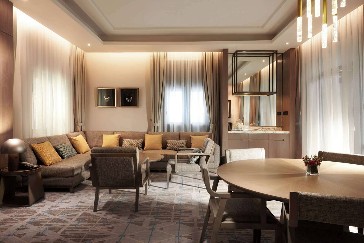 A modern hotel room with a sofa, dining table, chairs, and a kitchen area.