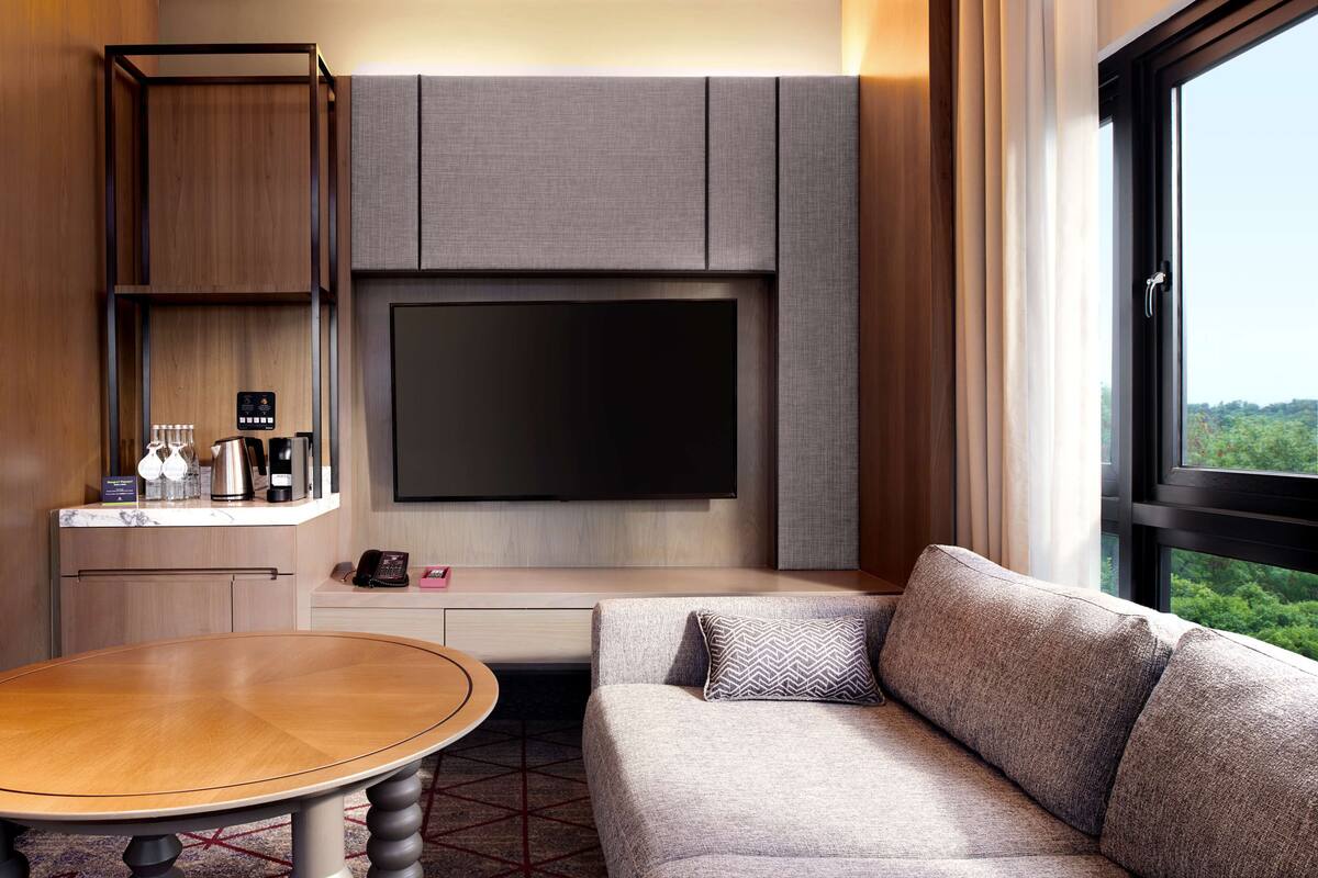 A modern hotel room with a large flat-screen TV mounted on the wall, a sofa, a small table, and a minibar.