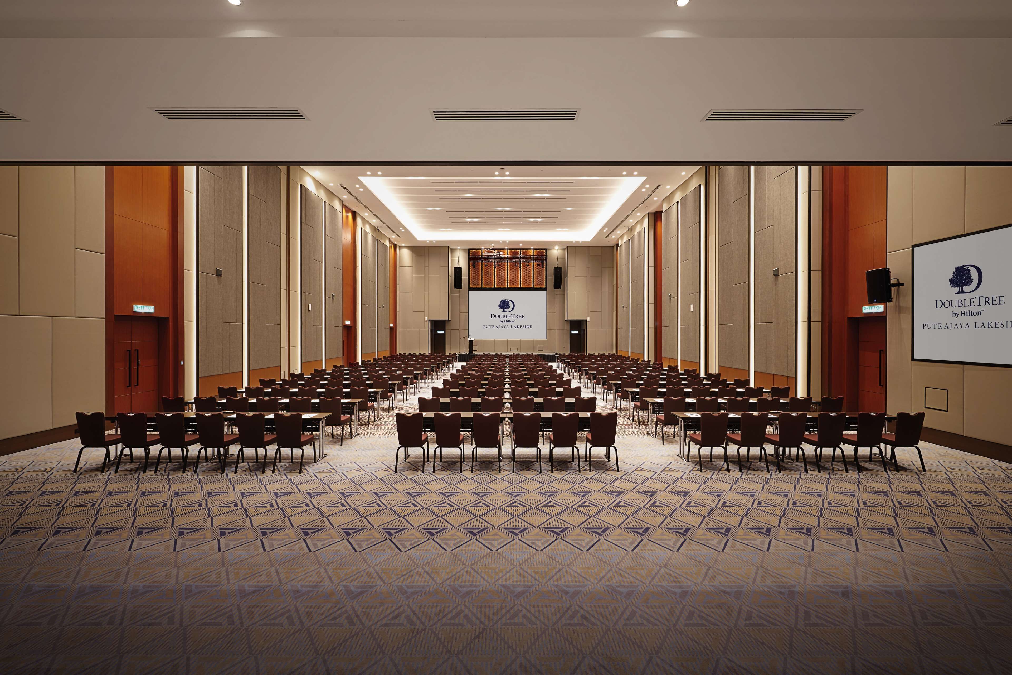 A conference room with rows of tables and chairs, a large screen at the front, and a carpeted floor.