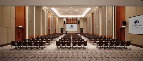 Un salón de conferencias con filas de mesas y sillas, una pantalla grande al frente y suelo alfombrado.