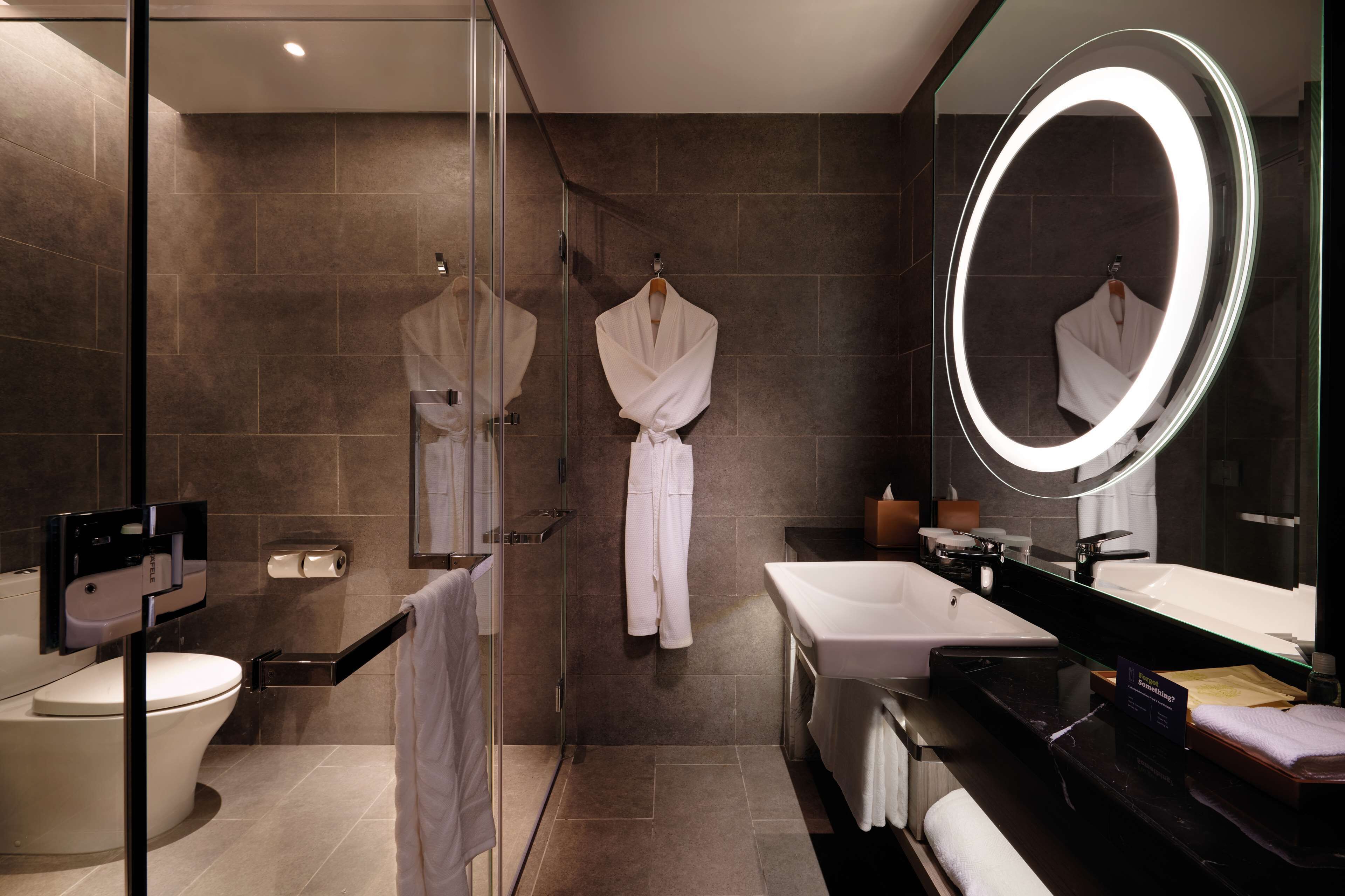 Une salle de bain moderne dotée d’une douche à parois de verre, de toilettes blanches, d’un grand miroir avec éclairage intégré et d’un meuble-lavabo avec un lavabo et un miroir.