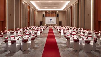 A banquet hall with rows of round tables, each with white tablecloths and red sashes, and a red carpet leading to a stage.
