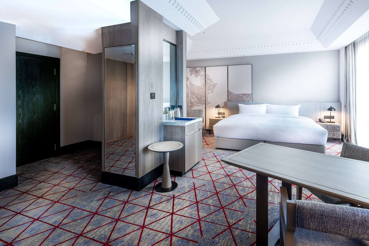 A hotel room with a bed, a desk, a chair, a small table, a mirror, a bathroom with a shower, and a patterned carpet.