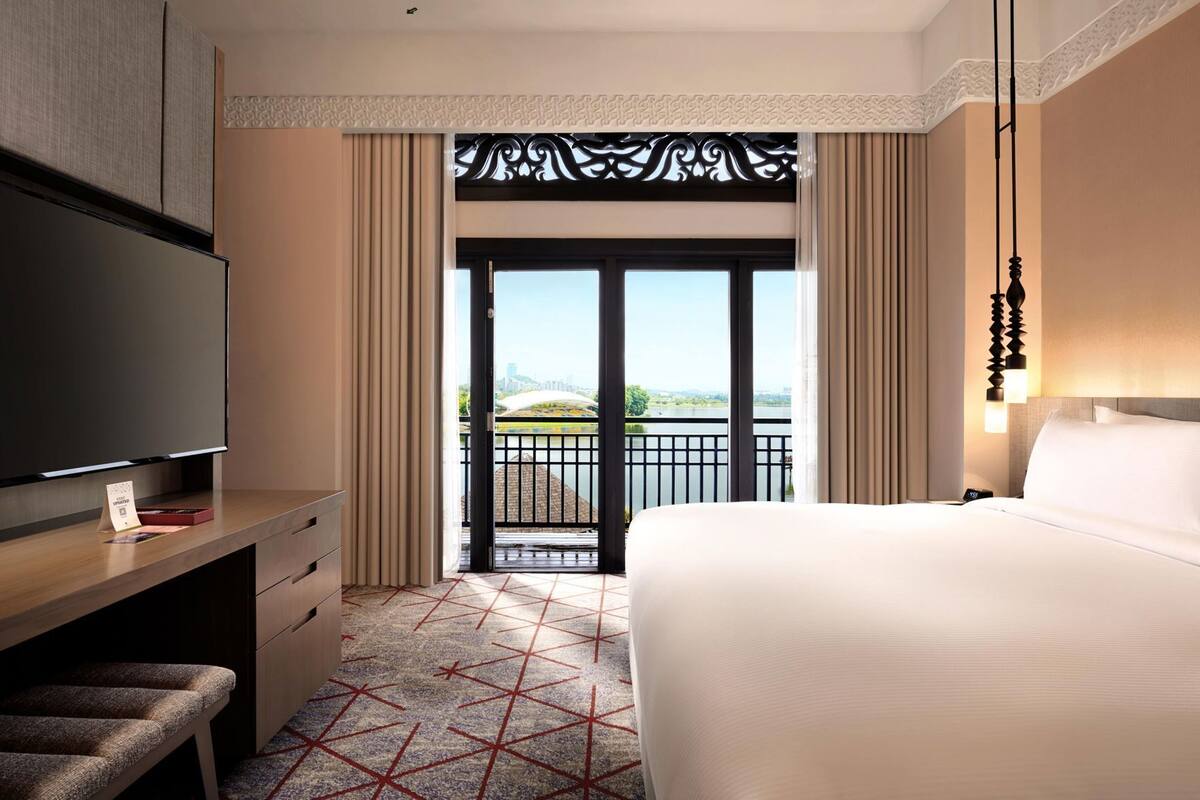 A hotel room with a large bed, a TV, a desk, and a balcony with a view of the city.