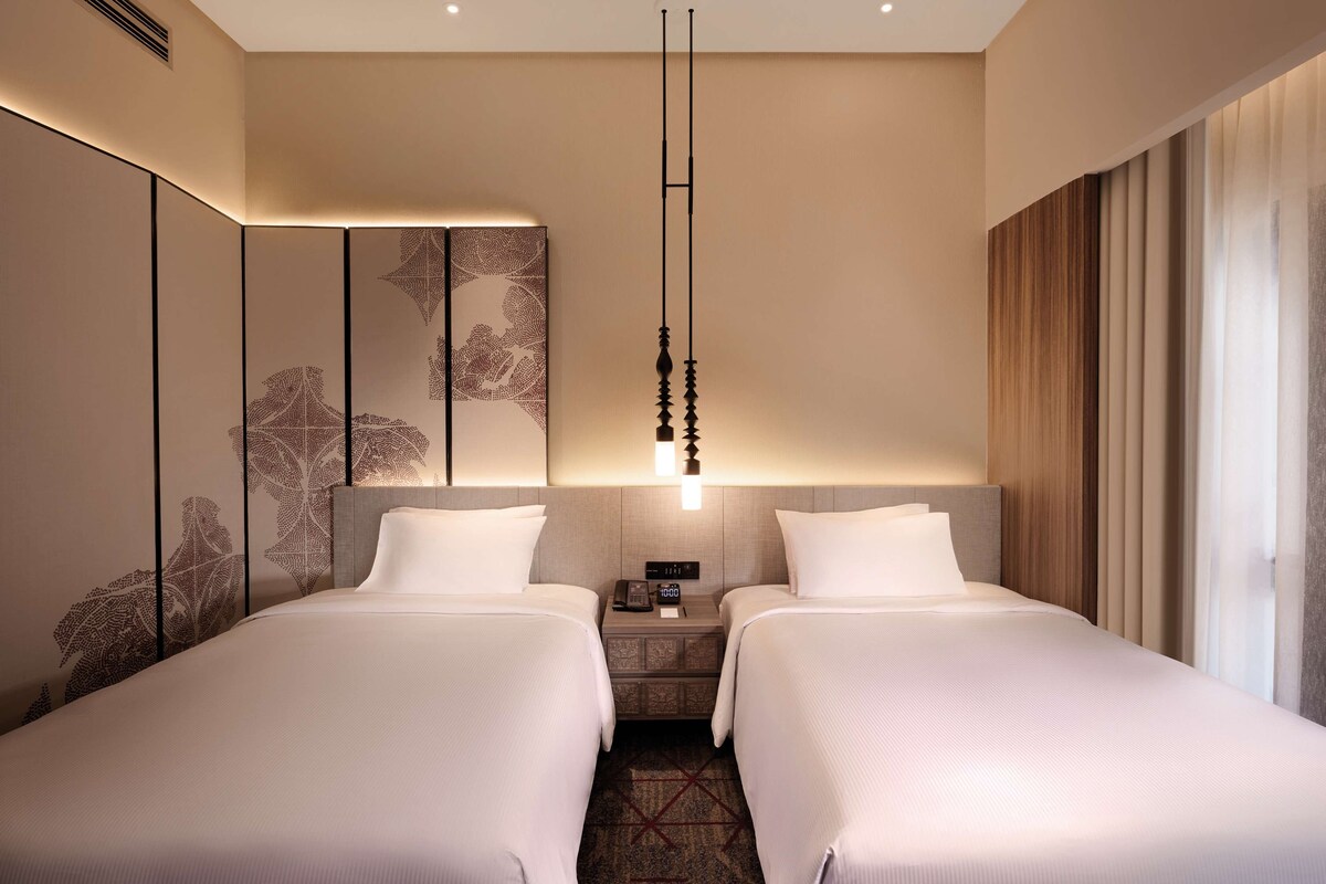 Habitación de hotel con dos camas, una mesita de noche con un reloj, una lámpara de pared y un ventanal con cortinas.