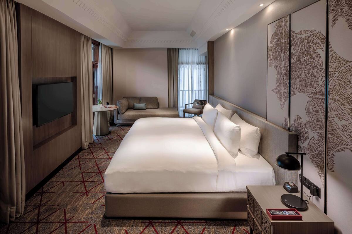 A modern hotel room with a large bed, a sofa, a TV, and a desk with a lamp.