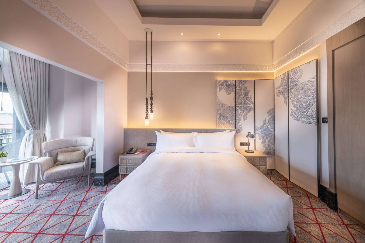 A hotel room with a large bed, a sitting area with a chair, a small table, a nightstand, and a large window with curtains.