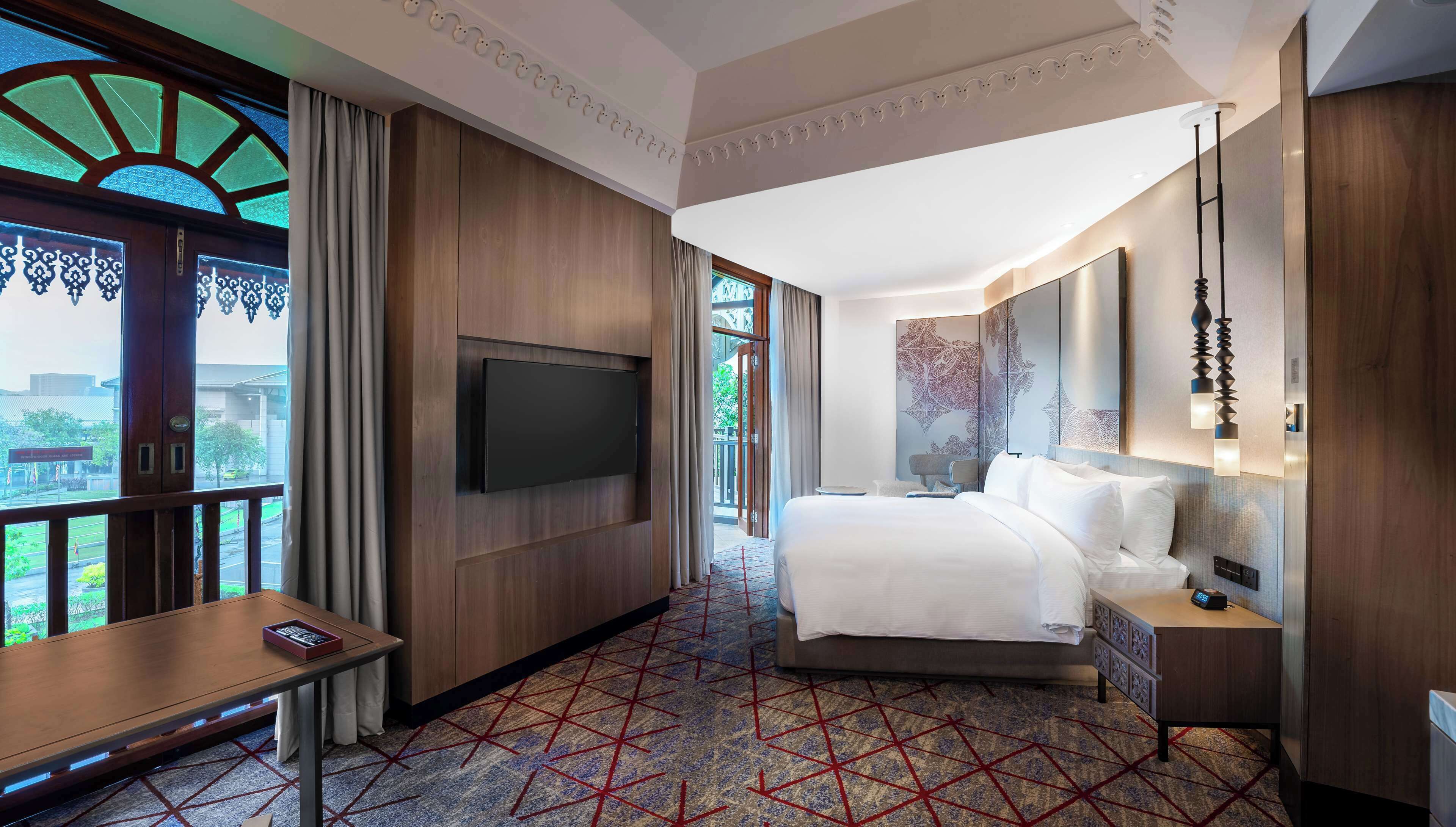 A hotel room with a bed, a TV mounted on the wall, a desk with a chair, and a view of the outside through a large window.