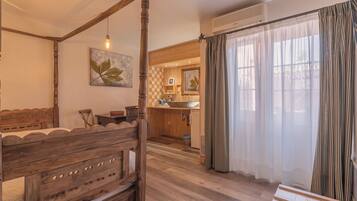 A bedroom with a wooden four-poster bed, a small table, and a chair. There is a kitchen area with a sink and a window with curtains.