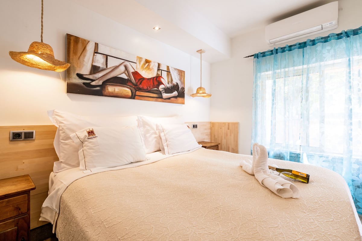 A hotel room with a large bed, white bedding, a painting on the wall, and a window with blue curtains.