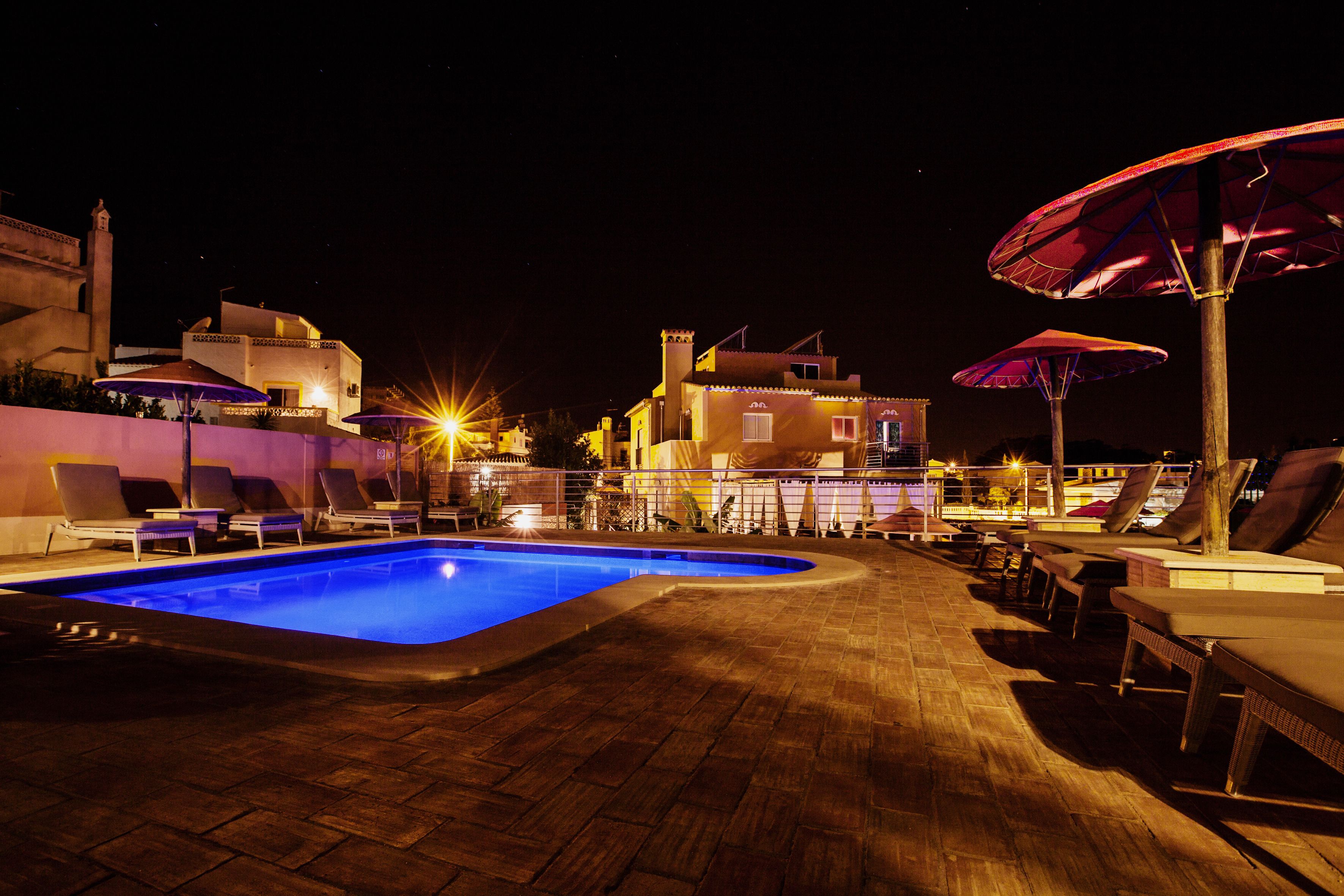 A swimming pool with lounge chairs and umbrellas, surrounded by buildings with warm lighting.