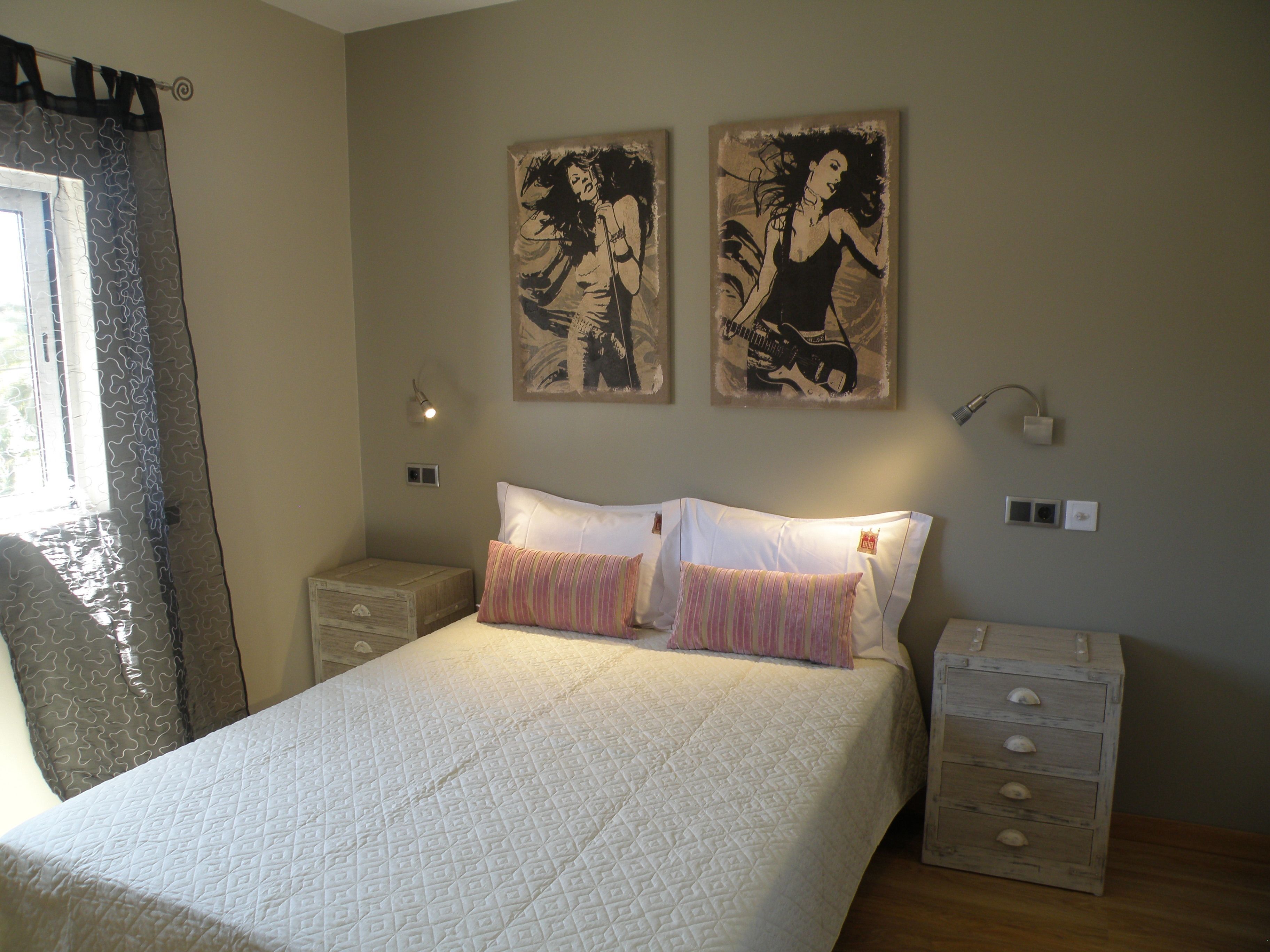 Een slaapkamer met een bed, twee kussens, een nachtkastje, een dressoir en twee aan de muur hangende schilderijen.