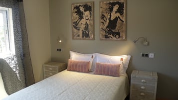 Een slaapkamer met een bed, twee kussens, een nachtkastje, een dressoir en twee aan de muur hangende schilderijen.