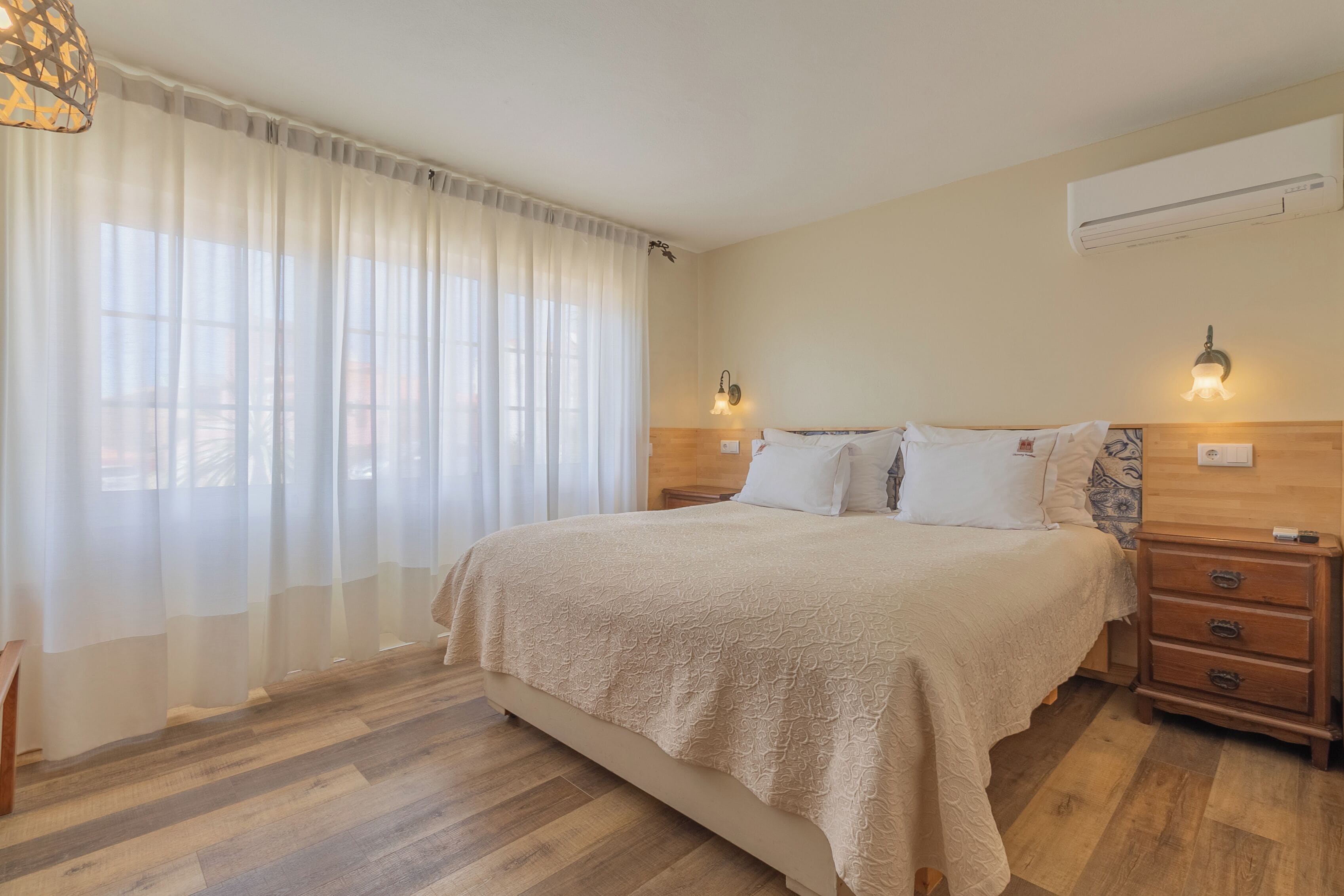 A bedroom with a large bed, wooden nightstands, a wall-mounted air conditioner, and sheer curtains.