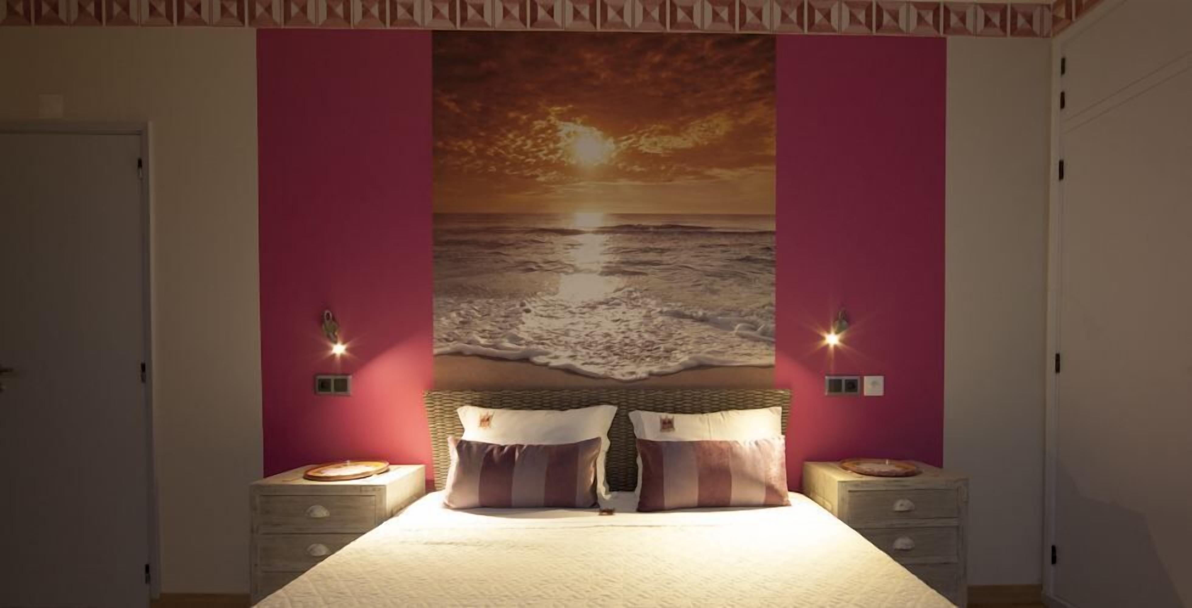 Une chambre à coucher avec un grand lit, deux tables de chevet et une fresque murale représentant un coucher de soleil sur l’océan.