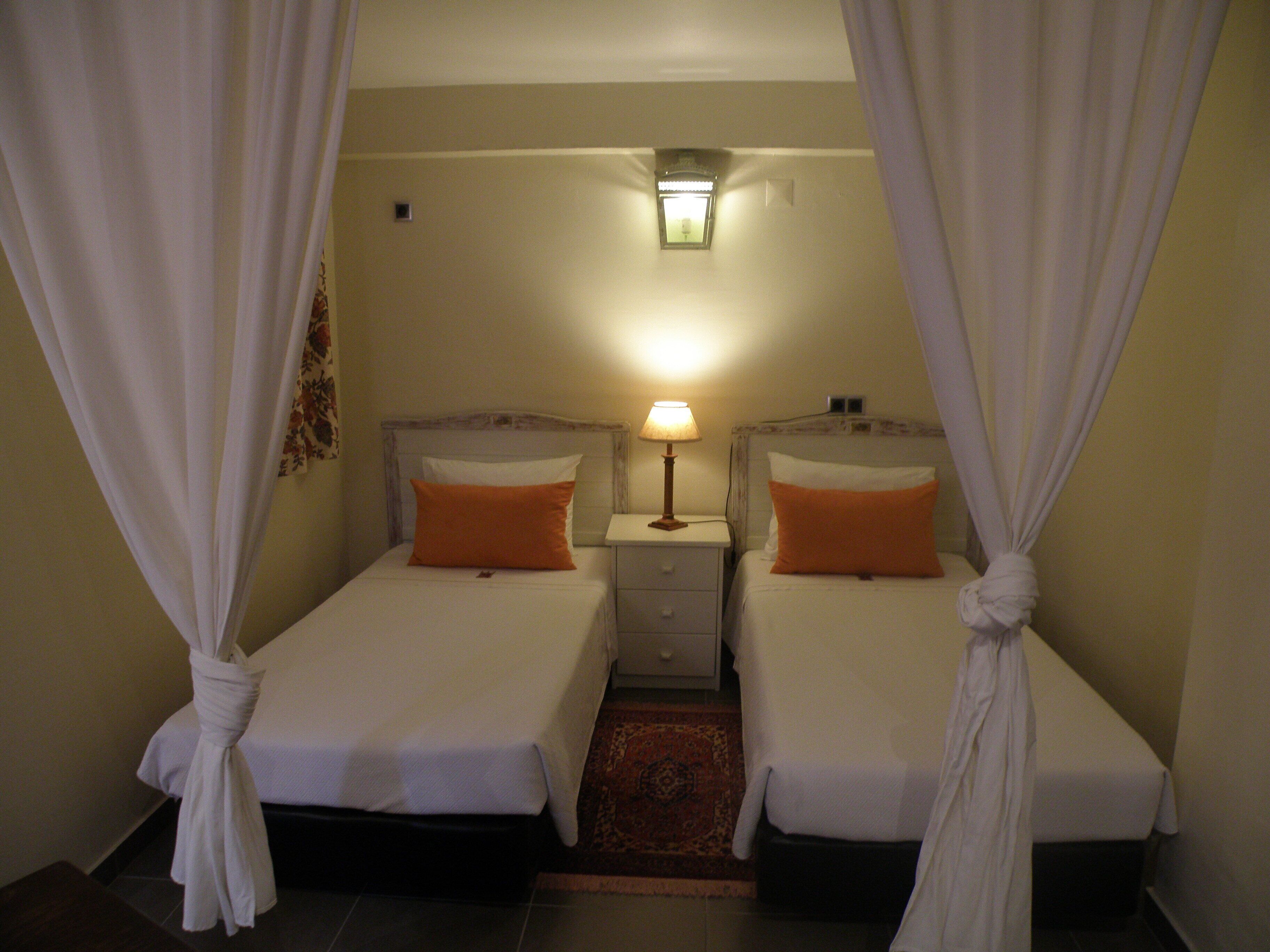 Habitación de hotel con dos camas individuales, cada una con un almohadón naranja y ropa de cama blanca. Hay una mesita de noche con una lámpara entre las camas. La habitación tiene una alfombra pequeña en el suelo y una lámpara de pared sobre la mesita de noche.