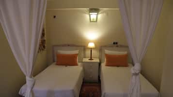 Habitación de hotel con dos camas individuales, cada una con un almohadón naranja y ropa de cama blanca. Hay una mesita de noche con una lámpara entre las camas. La habitación tiene una alfombra pequeña en el suelo y una lámpara de pared sobre la mesita de noche.