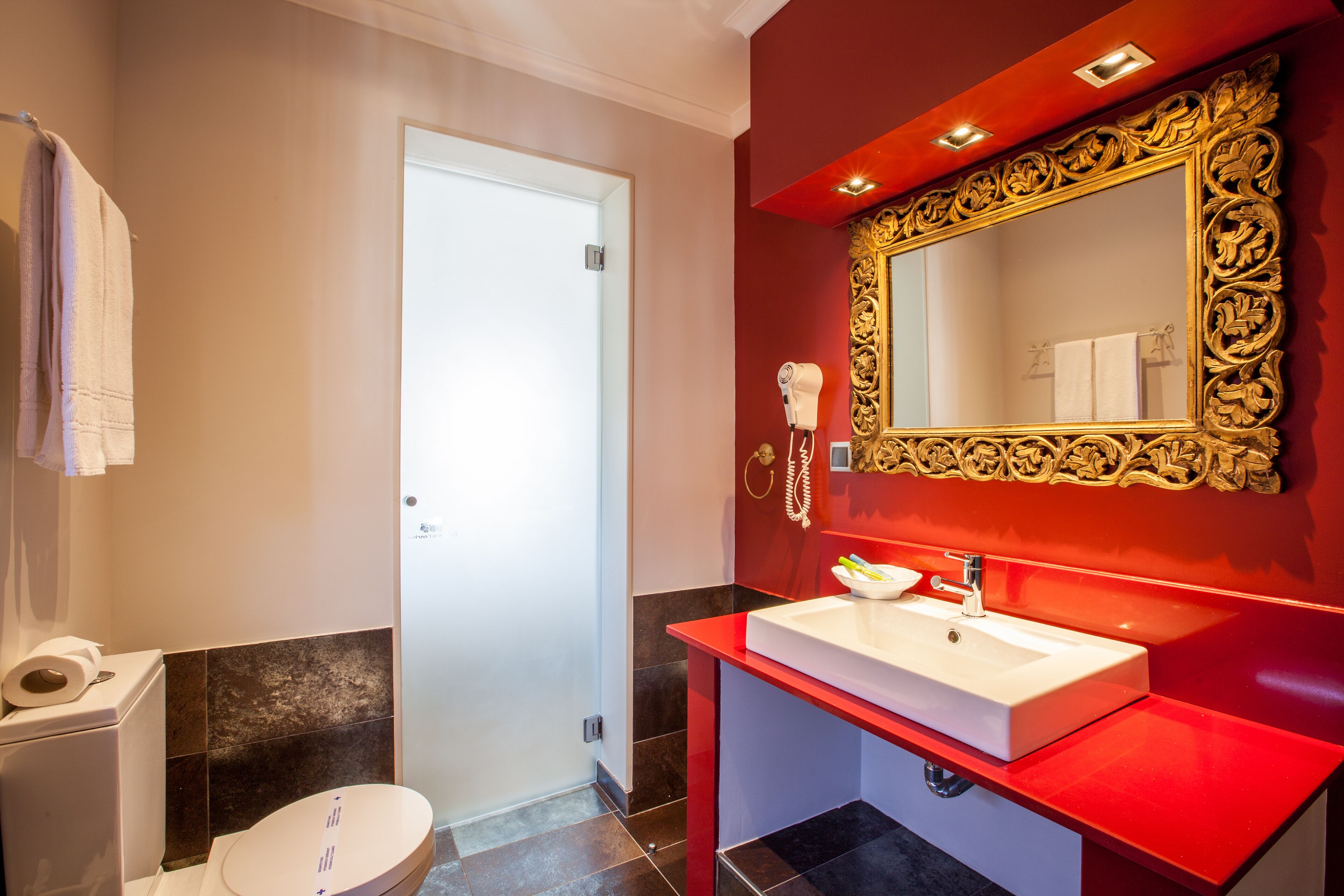 Un baño con inodoro blanco, tocador rojo con espejo de marco dorado y lavabo blanco.
