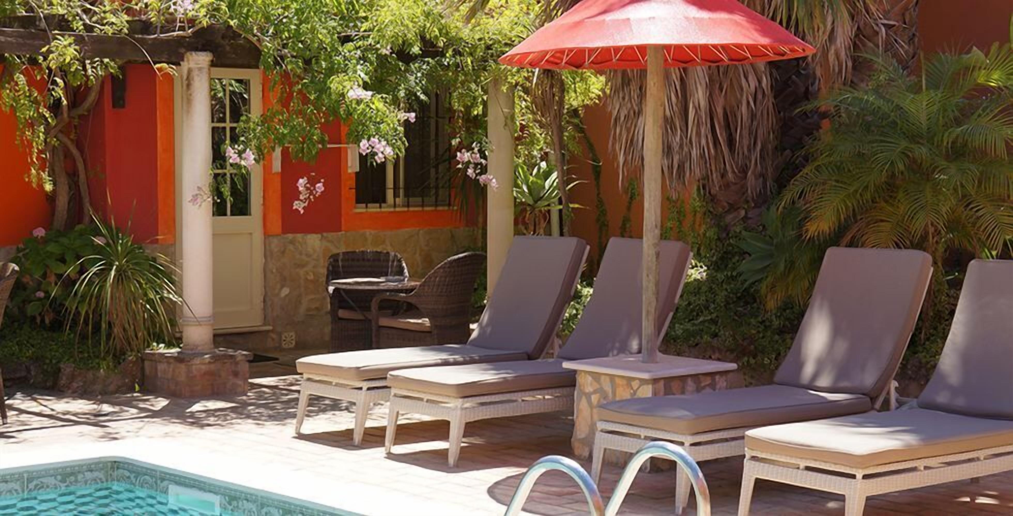 A poolside area with lounge chairs and a red umbrella, surrounded by greenery and a building with orange walls.