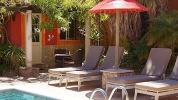 A poolside area with lounge chairs and a red umbrella, surrounded by greenery and a building with orange walls.