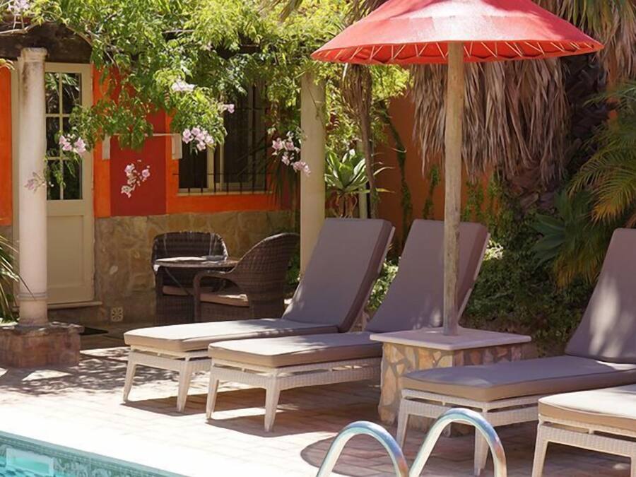 A poolside area with lounge chairs and a red umbrella, surrounded by greenery and a building with orange walls.