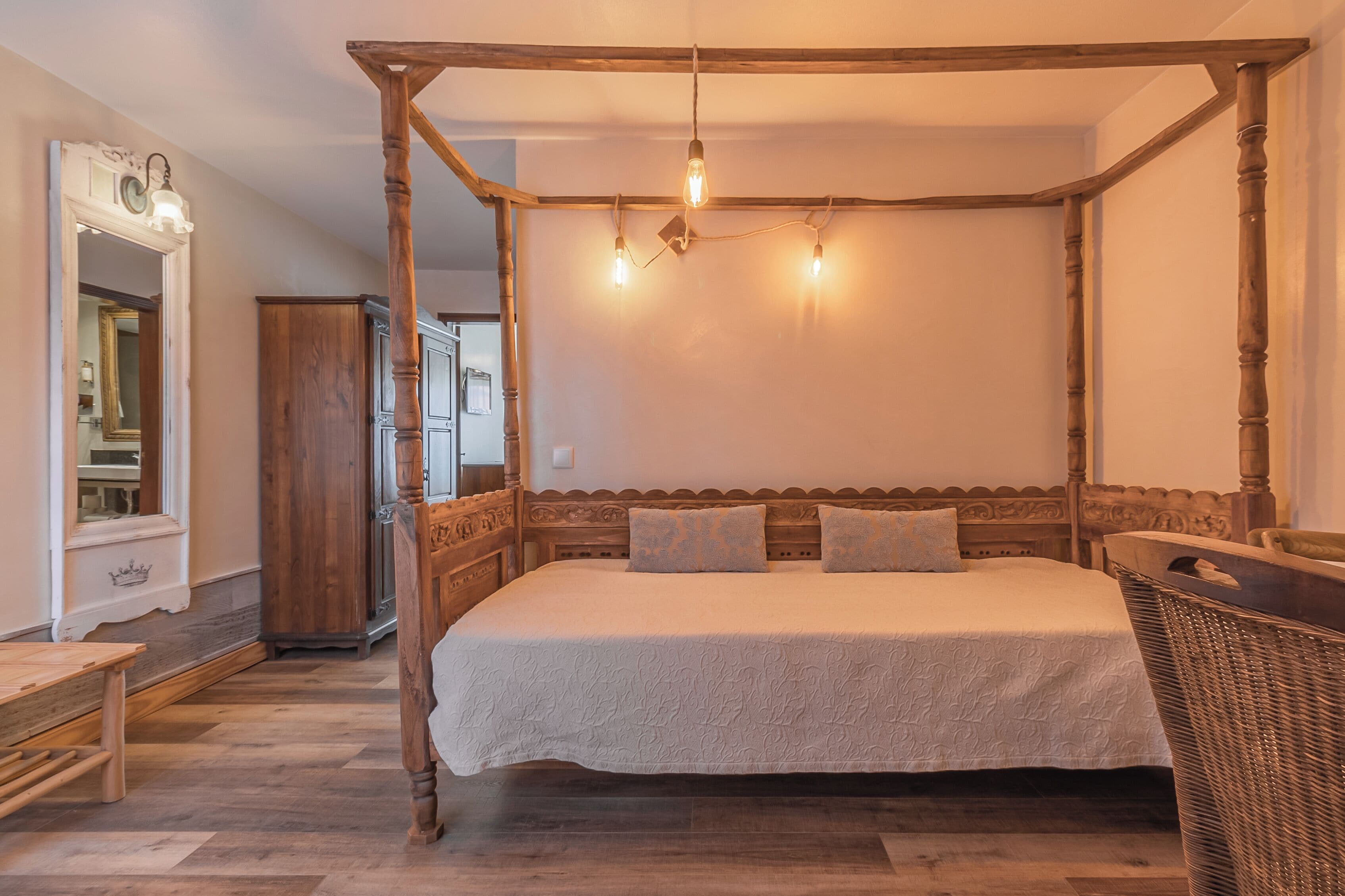 A four-poster bed with a wooden headboard, a bench, and a wicker chair in a room with wooden flooring and a mirror.