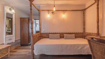 A four-poster bed with a wooden headboard, a bench, and a wicker chair in a room with wooden flooring and a mirror.
