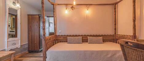 A four-poster bed with a wooden headboard, a bench, and a wicker chair in a room with wooden flooring and a mirror.